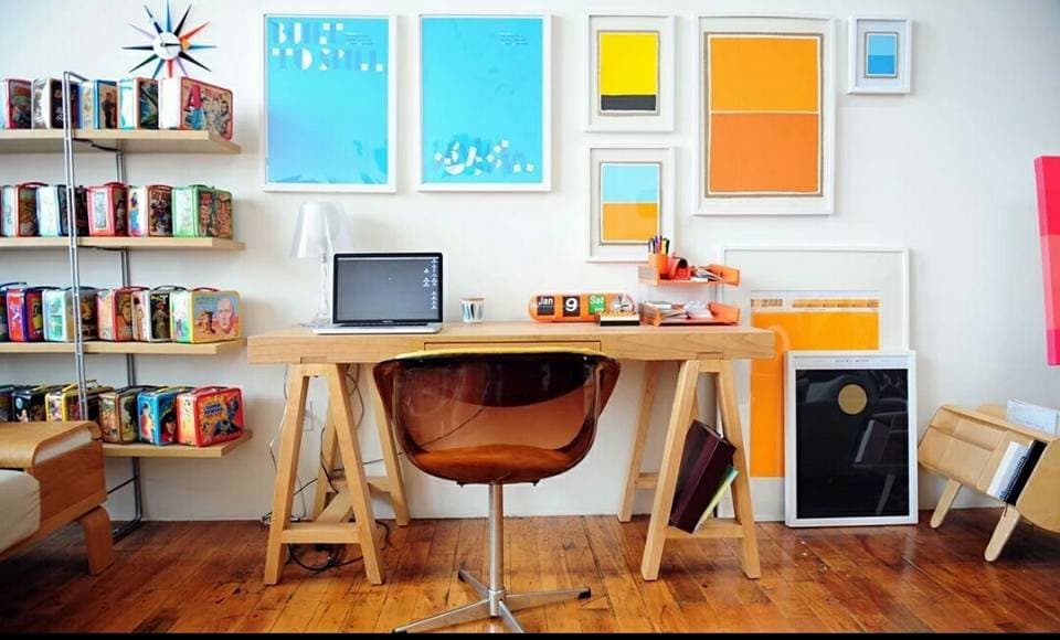 Contemporary-Wooden-Working-Desk-with-Cross-Legs-on-both-Sides-Acrylic-Brown-Swivel-Chair-surrounded-by-many-Wall-Framed-Paintings-creating-a-Eye-catching-Wall-Collage-in-a-Room. Design Detail Banner 0