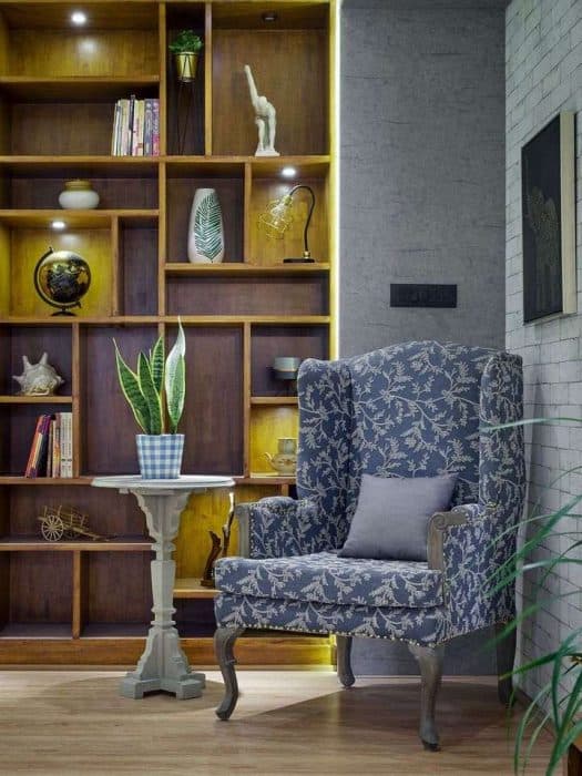 High-Wingback-Blue-Coloured-Floral-Chair-with-Wooden-Grey-Legs-Cushion-Pedestal-Circular-Side-Table-with-Snake-Potted-Plant-on-it-Back-side-is-Full-of-Storage-shelves Swiper Content 0