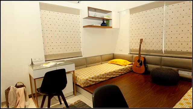Teens-Bedroom-with-Bed-Mattress-on-Raised-Level-with-Tufted-Headboard-on-all-3-sides-of-Wall-Wooden-Flooring-Pouf-Symmetrical-Working-Desks-with-Chairs-on-both-Sides-with-Shelves-on-Wall-2 Teens Bedroom with Bed